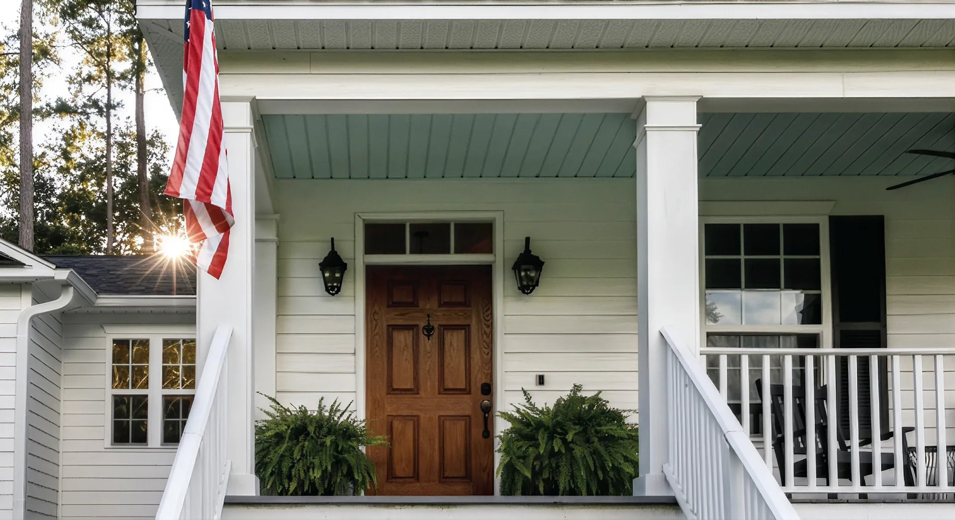 Covered front porch with a wooden entry door, hanging American flag, and white railing