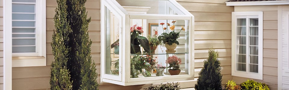 Garden window projecting from tan siding, its glass shelves full of potted flowers