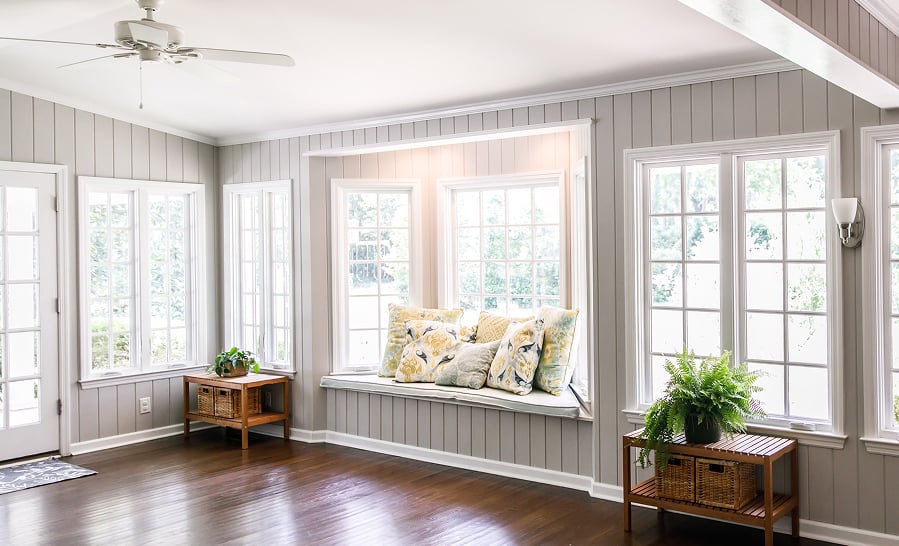 Sunroom lined with white double-hung and picture windows around a cushioned window seat