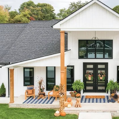 White farmhouse with black-framed windows, wooden porch posts, and autumn pumpkins on the front steps