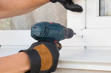 Gloved hands driving a screw into white window trim with a cordless drill