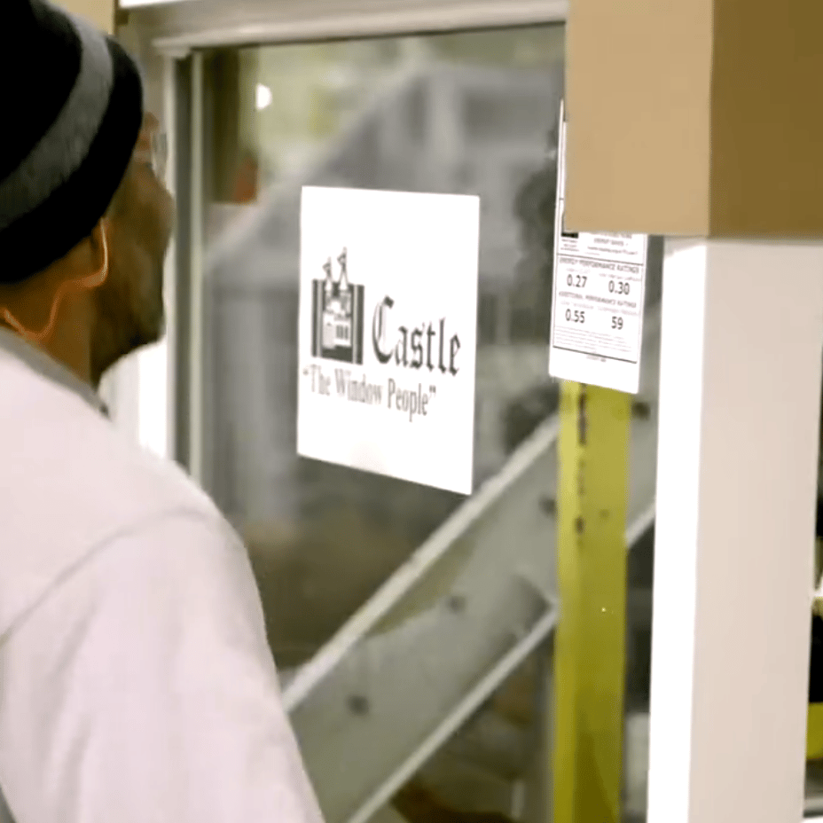 A man wearing a beanie stands near a window with a sign that reads 'Castle - The Window People.'