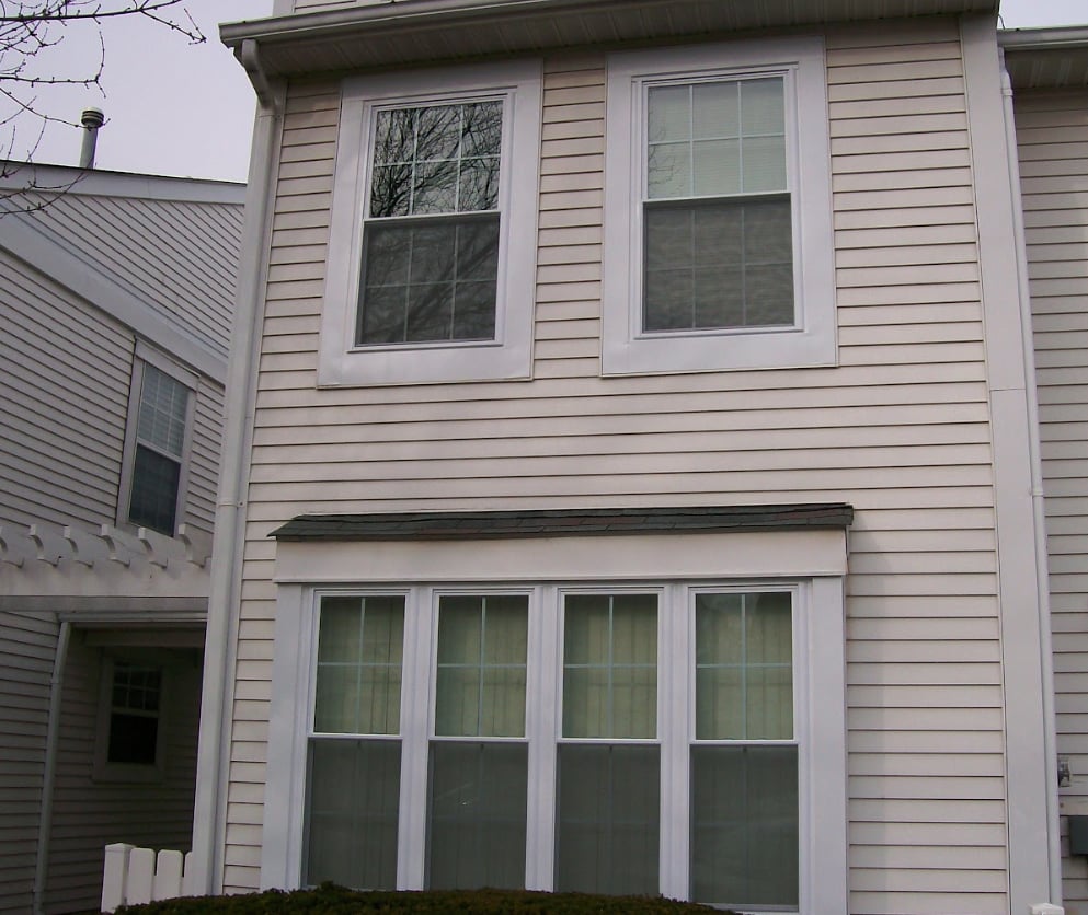 Exterior view of a two-story house with light beige horizontal siding, white trim, two second-floor double-hung windows, and a four-panel ground-floor window bay under a small shingled roof.