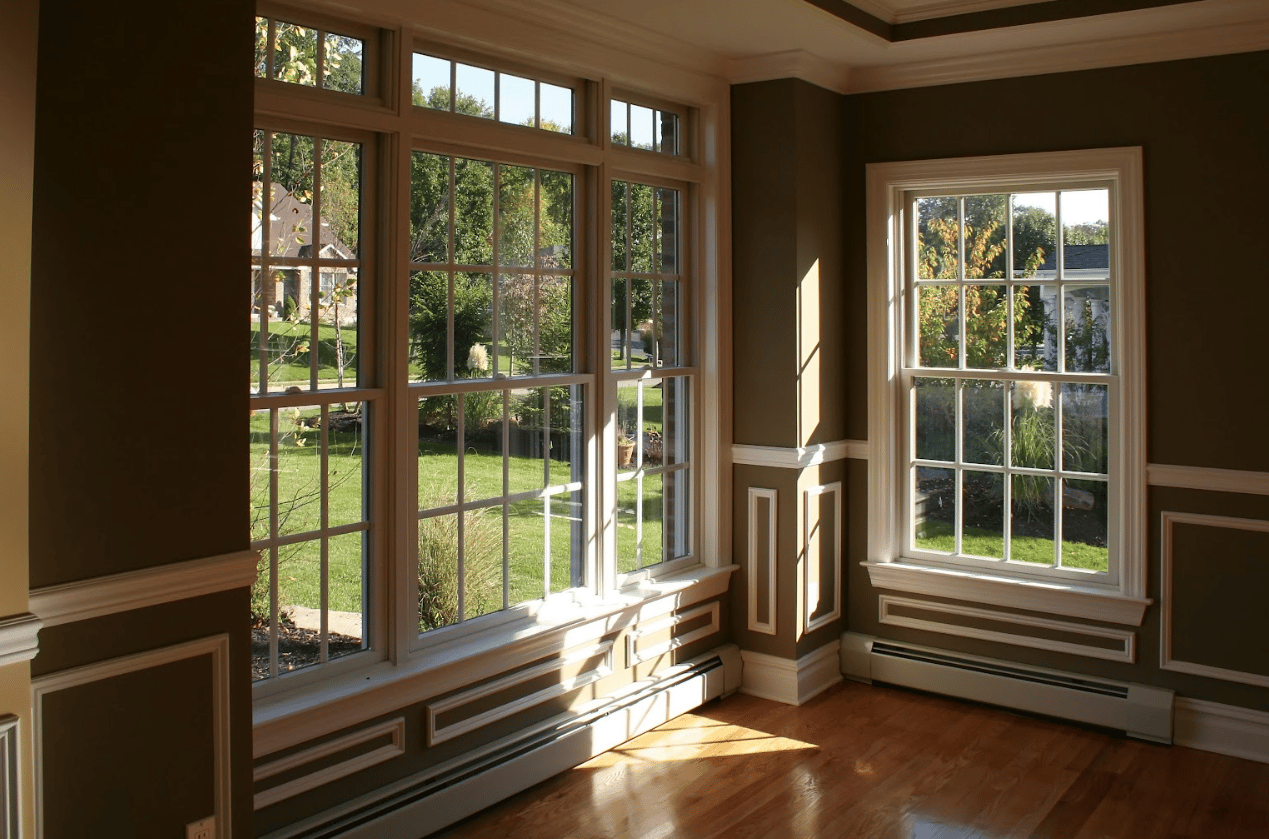Sunlit corner room with dark olive green walls, white wainscoting, hardwood floors, and large multi-pane windows looking out onto a grassy lawn.