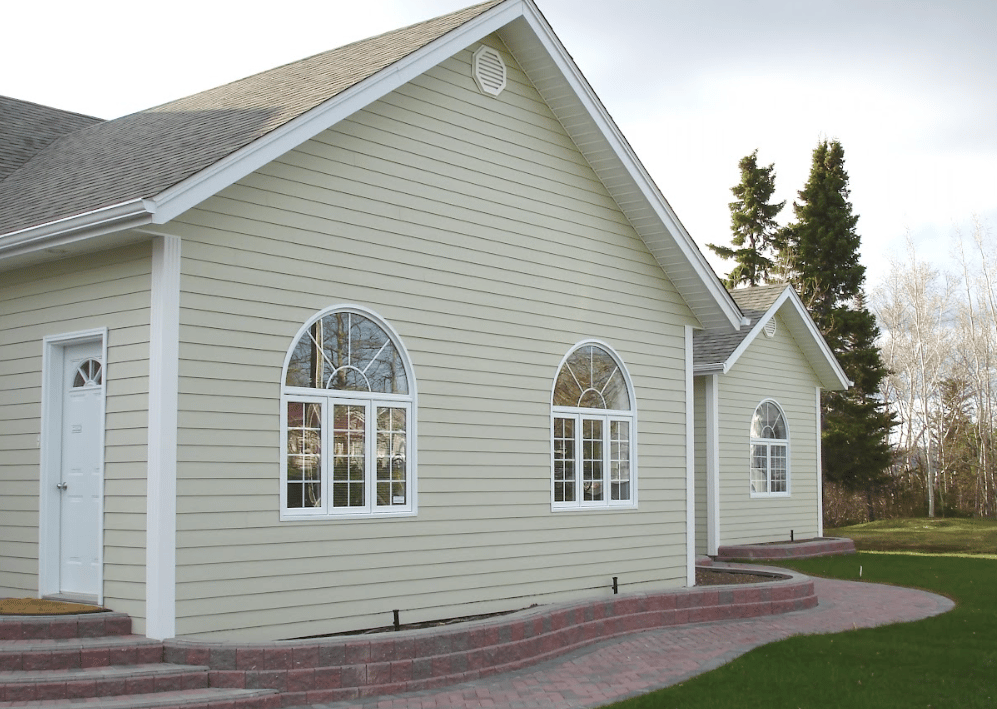 Exterior view of a light sage green house featuring white trim, arched sunburst windows, a side door, and a brick paver walkway bordered by a stacked stone garden bed.