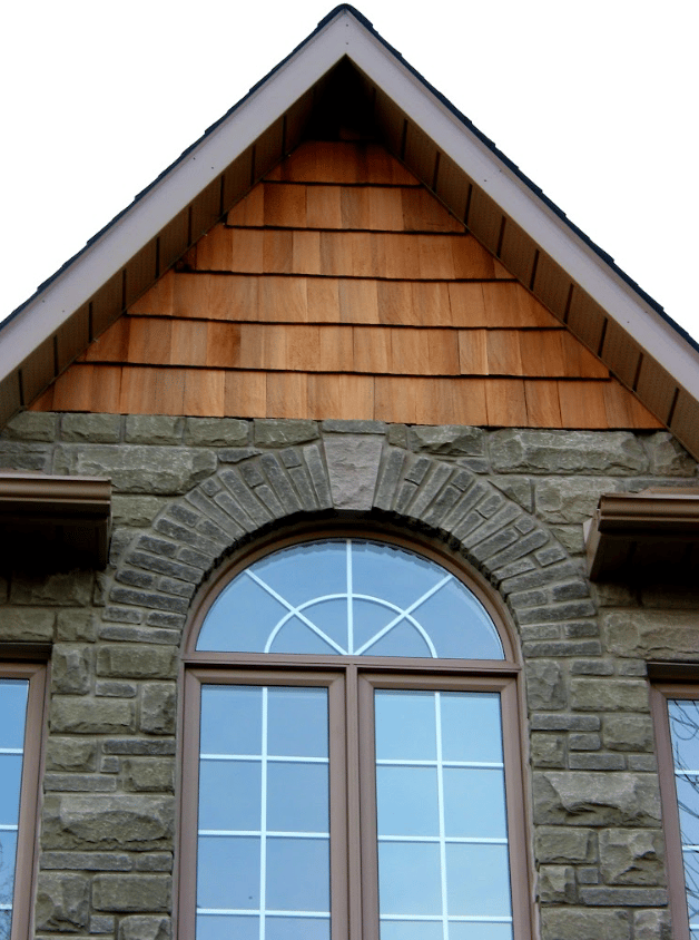 Exterior view of an arched window with light brown frames set into a stone facade, featuring a decorative stone arch above and a gabled roof peak with natural wood cedar shingles.
