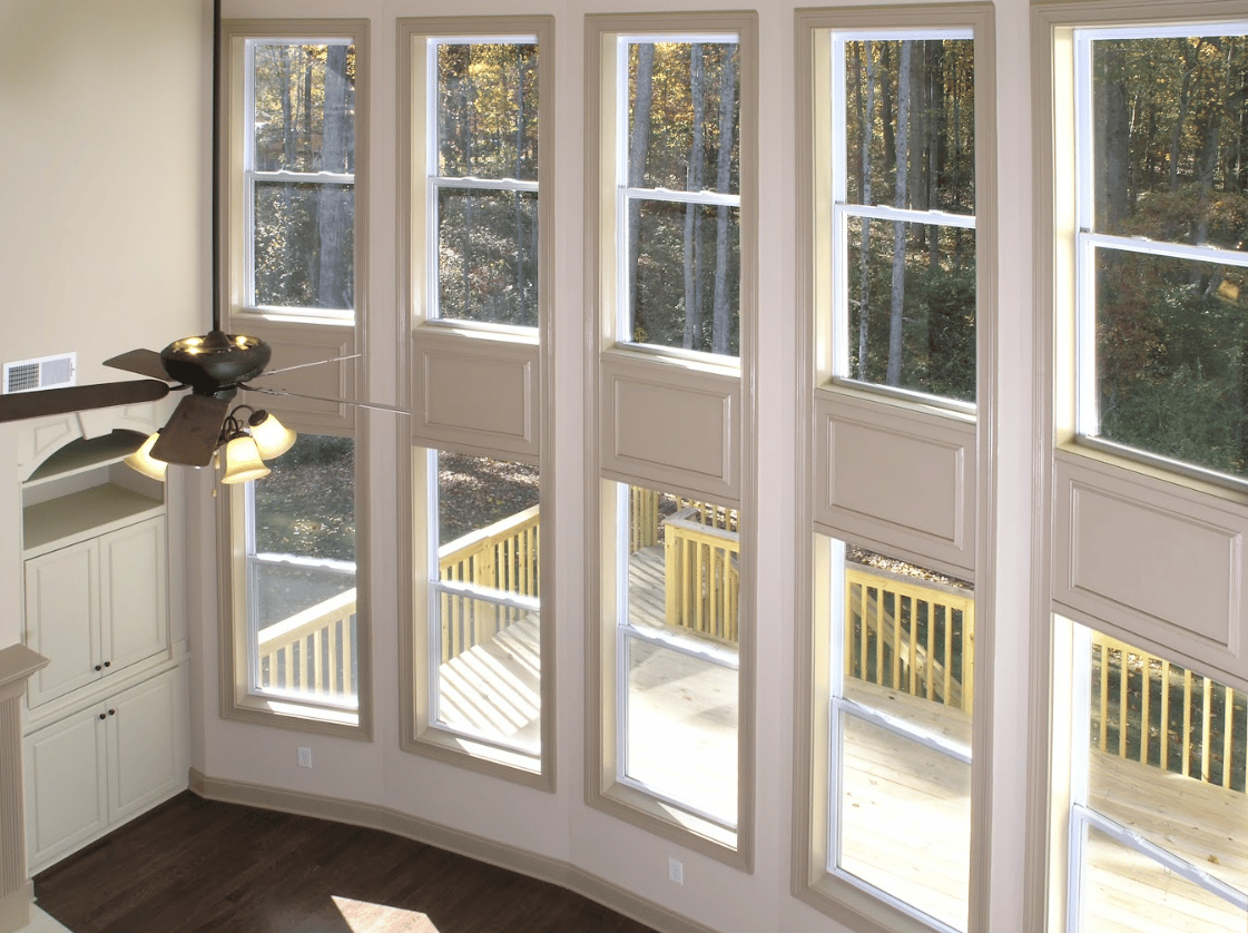 Interior high-angle view of a two-story living room featuring five floor-to-ceiling windows with beige trim, dark hardwood floors, a ceiling fan, and a view of a wooden deck and wooded yard outside.