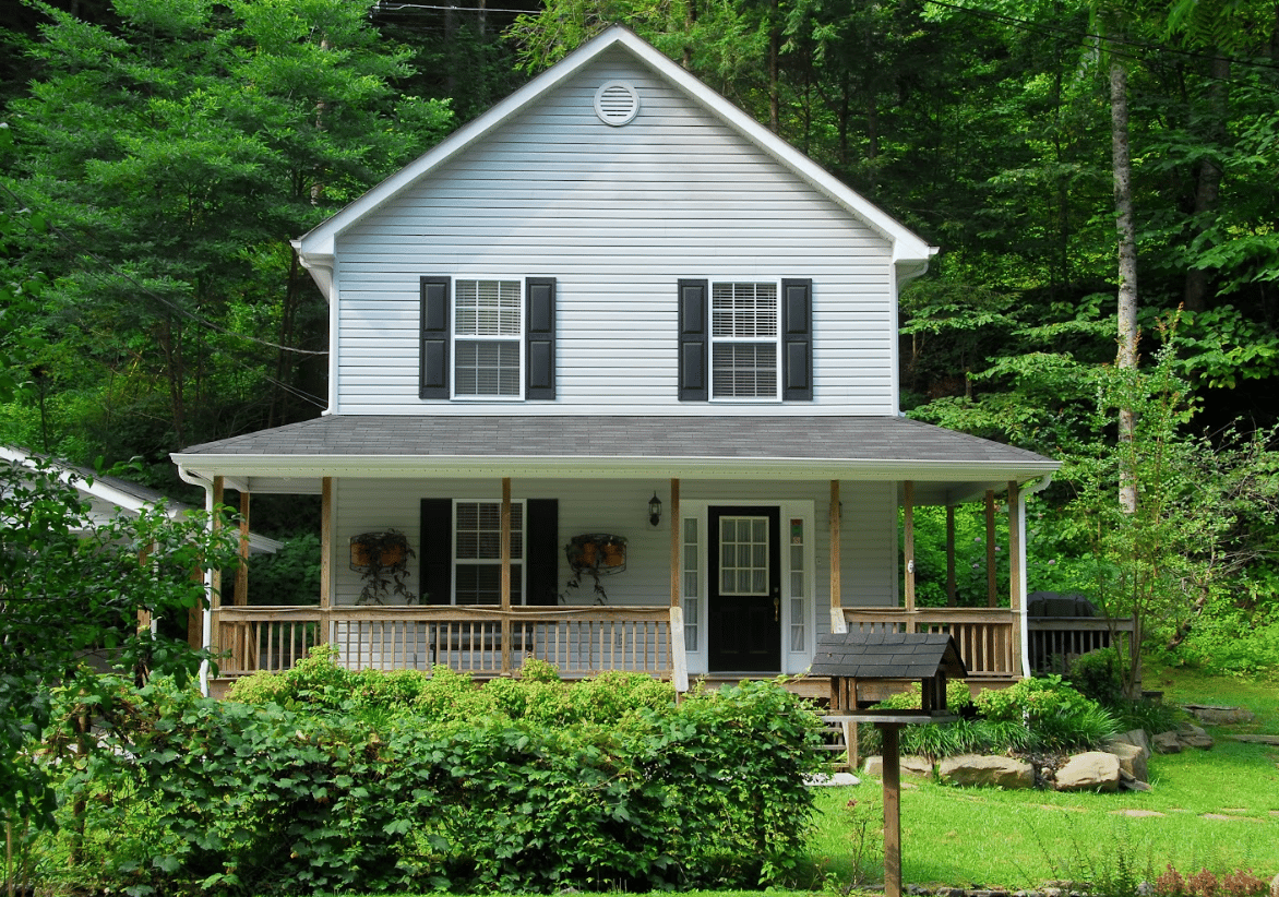Exterior view of a two-story light gray house with black shutters, a black front door, and a covered wooden front porch, nestled against a backdrop of dense green trees.