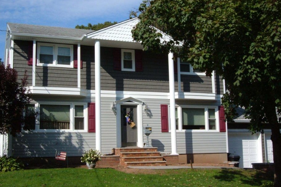 Exterior view of a two-story house featuring dark grey shake siding on the upper level, light grey horizontal siding on the lower level, red shutters, and white portico columns framing the front entrance.