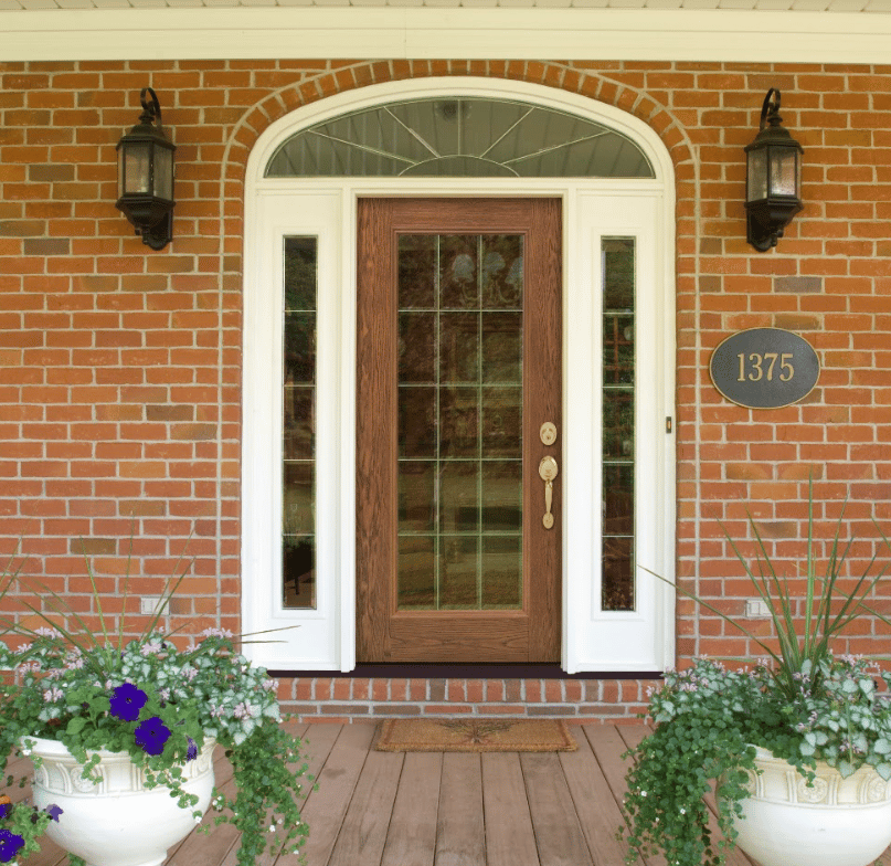 Exterior view of a forest green full-glass storm door set into tan horizontal siding, flanked by brass outdoor wall sconces and a white mailbox.