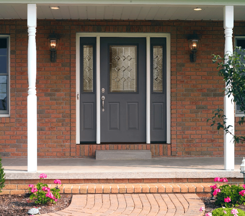 Exterior front entryway featuring a dark charcoal gray front door with decorative leaded glass inserts and matching sidelights, framed in white trim and set against a red brick wall with wall sconces.