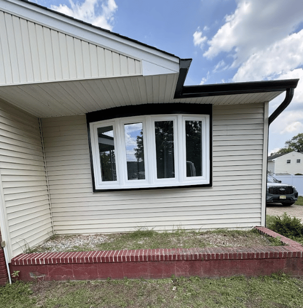 Exterior view of a bow window framed in dark trim on a light beige siding home, featuring a raised red brick garden planter along the front lawn.