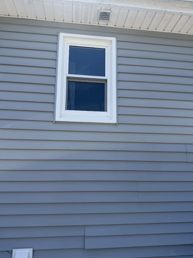 Exterior view of a single white-framed double-hung window set into light blue-gray horizontal vinyl siding below a white soffit.