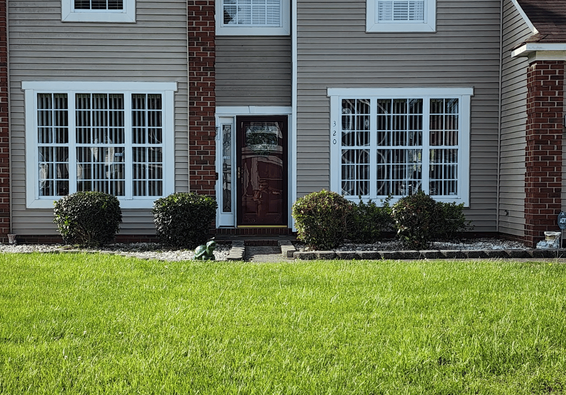 Exterior front view of a house with grey horizontal siding, red brick pillars, a dark front door, large white multi-pane windows, and manicured shrubs lining the lawn.