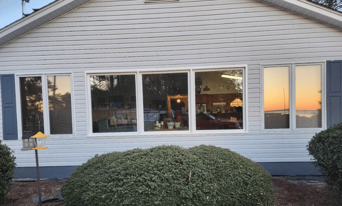 Exterior view of a single-story house with light grey siding, dark blue shutters, a series of large picture windows reflecting the warm glow of sunset, and dense green bushes in the foreground lawn.