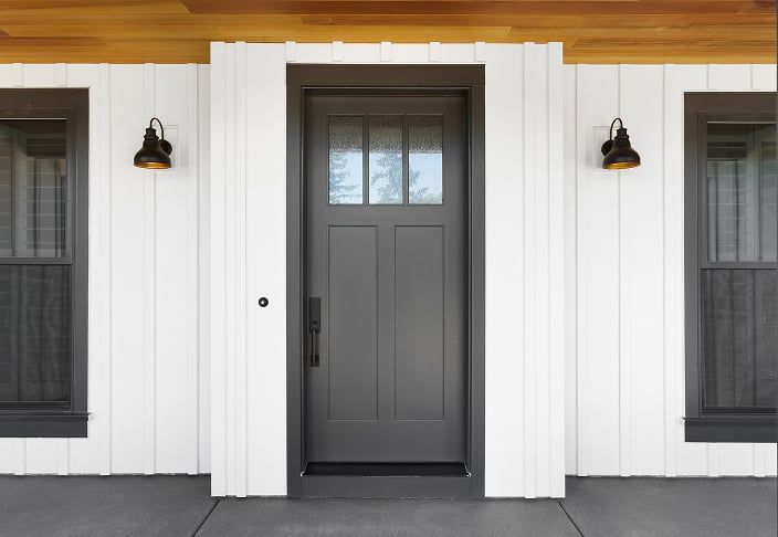 Dark gray custom entry door with a three-pane glass top, flanked by black sconces on white board-and-batten siding