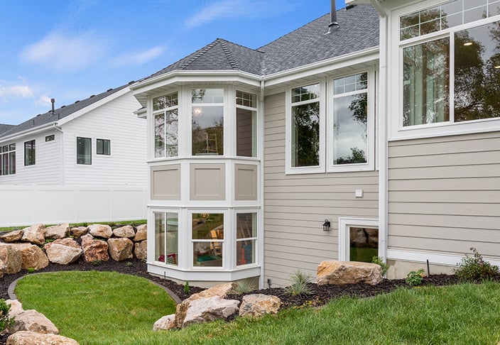 Angled bay window projecting from the tan siding of a home, with stone landscaping along the foundation